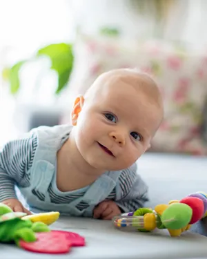 Baby smiling while lying on their stomach, surrounded by soft, colorful toys in a cozy room.