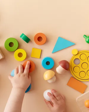 Baby's hands playing with colorful educational wooden blocks and geometric shapes on a light beige surface.