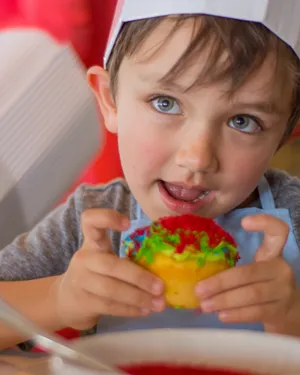 A young boy wearing a chef's hat happily holding a colorful, decorated cupcake.