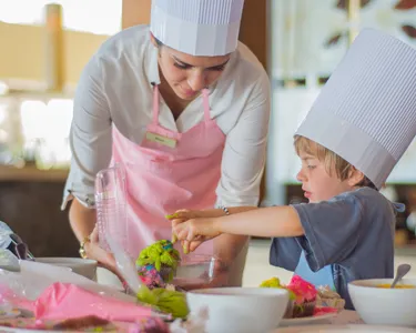 A chef helping a child decorate colorful food at a table, both wearing chef hats.