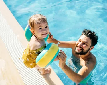 A father playing with his young child at the edge of a swimming pool, the child wearing floatation armbands and smiling.