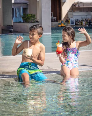 Two children sitting on the edge of a pool, enjoying colorful drinks while splashing in the water.