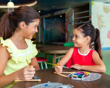 Two young girls sitting at a table, happily painting and doing crafts together indoors.