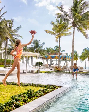 A young girl standing by a pool, preparing to throw a football, while a boy plays in the water in the background.