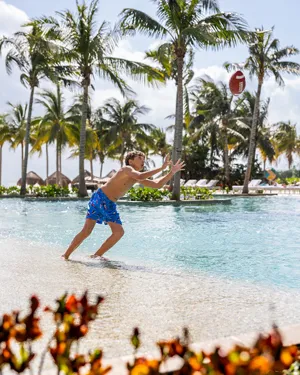 A boy running on the edge of a pool, reaching to catch a ball, with palm trees in the background.