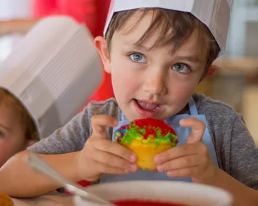 A young boy wearing a chef's hat, excitedly holding and tasting a colorful cupcake he decorated.